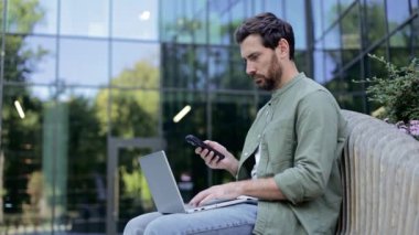 Bir iş adamı şirket binasının dışındaki bankta oturuyor, bilgisayarını ve cep telefonunu kullanarak uzaktan çalışıyor..
