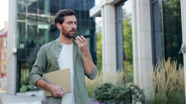 Genç bir işadamı bilgisayarını tutarken ve yürürken akıllı telefonuyla ses asistanını kullanıyor. Gülümsüyor ve mutlu görünüyor..