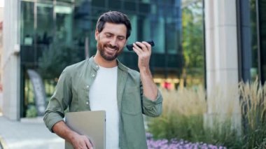 Genç, mutlu ve kendinden emin bir işadamı şehirde dizüstü bilgisayarıyla yürüyor ve telefonuyla konuşuyor. Gününe devam ederken başarılı ve mutlu görünüyor..
