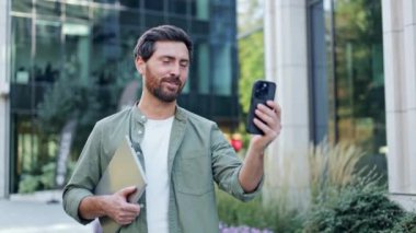 Genç, mutlu ve çekici bir iş adamı yürürken bir video görüşmesinde. Bir elinde telefonunu, diğer elinde laptopunu tutuyor..