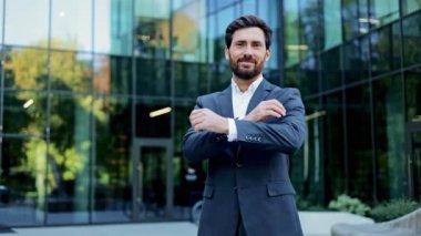 Takım elbiseli başarılı bir işadamı modern cam ofis binasının önünde kollarını kavuşturarak kameraya gülümsüyor..