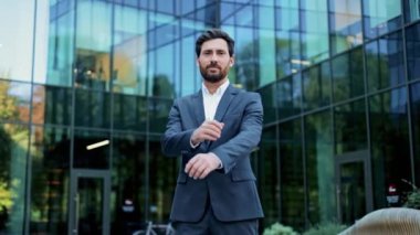 Takım elbiseli bir işadamı modern bir ofis binasının önünde duruyor. Kollarını kavuşturup kameraya gülümsüyor, mutlu ve başarılı görünüyor..