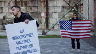 Liman işçileri otomasyon ve robotlar yüzünden iş kaybını protesto ediyorlar. İşçi hakları ve iş güvenliği için verdikleri mücadeleyi simgeleyen işaretler ve Amerikan bayrağı taşıyorlar..