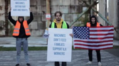Liman işçileri otomasyon ve robotlar yüzünden iş kaybını protesto ediyorlar. Gençlerin elinde, işlerini ve haklarını geri isteyen bir Amerikan bayrağı ve tabelaları var. Bir işçi megafona konuşuyor..