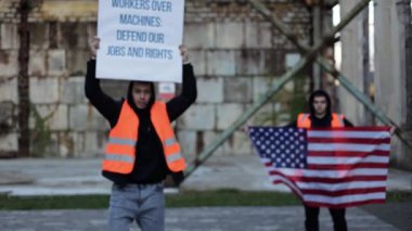 Otomasyon ve iş kaybını protesto eden iki Amerikalı işçi bir depoda pankart ve bayraklar tutuyor..