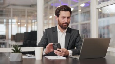 Modern bir ofisteki masasında oturan bir işadamı, telefonunu ve bilgisayarını kullanıyor. İşine odaklanmış ve mesajlarını kontrol ediyor..