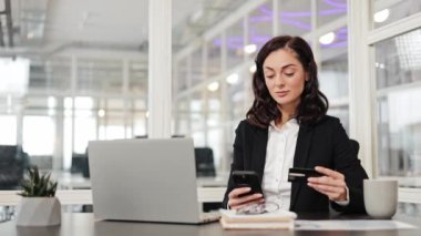 Ofisteki iş kadını akıllı telefon ve kredi kartı kullanarak online alışveriş yapıyor. Kredi kartı tutan ve modern ofiste akıllı telefon kullanan kendine güvenen bir kadın..