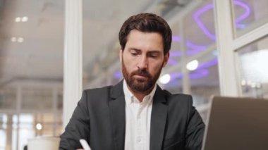 Bir iş adamı bir kafede dizüstü bilgisayarının önünde oturup not defterine bir şeyler yazıyor. Takım elbise, gömlek ve sakalı var. Kafe cam bölmeleri ve mor neon ışıklarıyla modern..