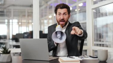 Kızgın işadamı megafona bağırıyor ve kamerayı işaret ediyor. İş adamı dizüstü bilgisayarla masada oturuyor. Ofis ayarlama. Stresli çalışma ortamı.