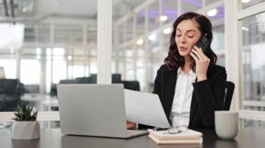 A businesswoman in a black suit sits at her desk and reviews a document while talking on her cell phone. The modern office is seen in the background.