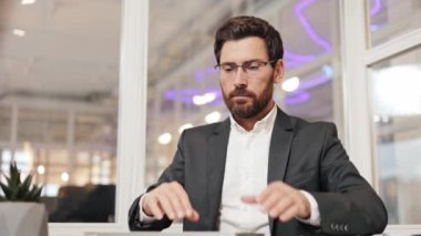 Takım elbiseli bir işadamı masasında oturur ve dizüstü bilgisayarı üzerinde çalışmak için gözlüklerini takar. Odaklanmış ve üretken..