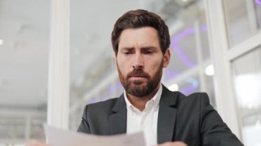 Takım elbiseli endişeli işadamı hayal kırıklığı ve endişe gösteren bir belge tutuyor. Adam, kurumsal bir ortamda kaygı ve stresi ifade ederek şakaklarını ovuyor..