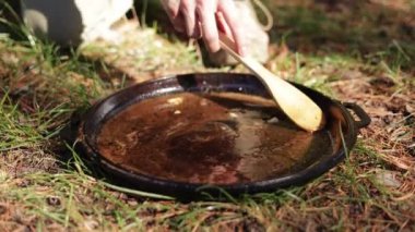 Koyu bir sıvıyı ahşap bir kaşıkla karıştıran birinin yakın çekimi. Tava çimlerin üzerinde, ilkel bir açık hava yemek sahnesini çağrıştırıyor..