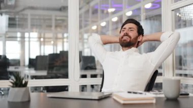 Mutlu iş adamı, elleri başının arkasında, ofis koltuğunda dinleniyor, işe ara veriyor. Gülümsüyor ve bir anlık huzurun tadını çıkarıyor. İş yeri sağlığı ve iş videoları için mükemmel..