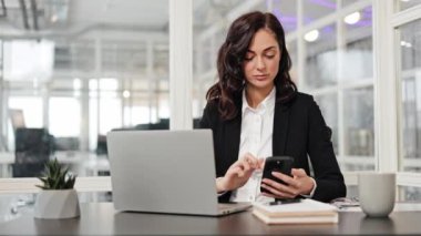 Yönetici iş kadını masa başında oturuyor, telefon ve dizüstü bilgisayar kullanıyor. Kadınlar modern ofis alanlarında odaklanmış ve üretkendir. Gülümseyen bir kadın iş yerindeki masada oturuyor ya da cep telefonunda mesaj yazıyor..