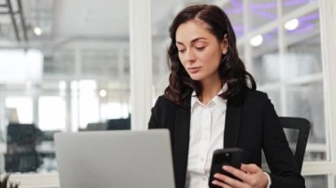 Neşeli iş kadını modern ofis masasında elinde dizüstü bilgisayarla kameraya gülümsüyor. Mesaj yazan, sohbet eden, sosyal medyayı karıştıran, e-postaları kontrol eden bir kadın..