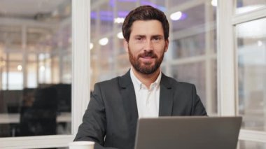 Takım elbiseli, gülümseyen mutlu iş adamı ofisinde oturuyor ve kameraya gülümsüyor. İdari lider, müdür kendinden emin, başarılı ve dost canlısı görünüyor..