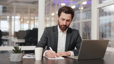 Ofiste dizüstü bilgisayarla çalışırken not defterine notlar yazan bir iş adamı. İş, ofis, iş, bilgisayar, dizüstü bilgisayar, defter, yazı, notlar, iş adamı, adam, takım elbise.