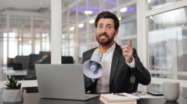 Takım elbiseli bir işadamı megafondan önemli bir duyuru yapıyor, modern cam ofisindeki kameraya bakıyor. Laptop, not defteri, masadaki akıllı telefon. Doğal ışıklandırma.