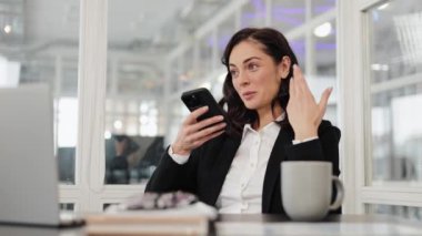 Takım elbiseli gülümseyen iş kadını ofisteki masasında oturup konferans görüşmesi için telefonunu kullanıyor. Laptopuna bakıyor ve gülümsüyor..
