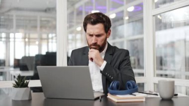 Odaklanmış işadamı, modern ofis ortamında dizüstü bilgisayarı üzerinde çalışıyor. Yüzündeki düşünceli ifade derin bir konsantrasyon ve işine bağlı olduğunu gösteriyor..