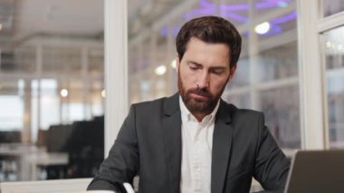 Takım elbiseli bir işadamı ofisinde masasında oturuyor ve kameraya gülümsüyor. Mutlu ve kendinden emin görünüyor. İş adamı, girişimci ya da erkek çalışan. İş yerindeki mutlu bir ofis çalışanının yüzü