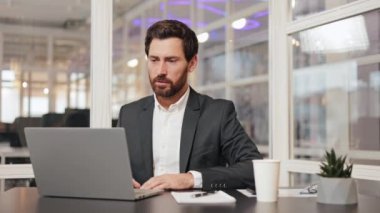 Ciddi bir iş adamı ofisinde oturuyor ve bilgisayarını kullanarak video konferansı yapıyor. İş adamı çağrısı, çevrimiçi konferans, ofisteki bilgisayar ekranına bakmak, uzaktan iş eğitimi.