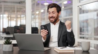 İş adamı ofiste telefon ve dizüstü bilgisayar kullanırken başarıyı kutluyor. İnsanoğlu başarının sevincini ve heyecanını ifade eder. Mobil galibiyet, heyecan dolu sevinç dolu şanslı yönetici iyi haberler alıyor..