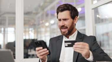 Takım elbiseli bir işadamı çevrimiçi ödemeyle ilgili sorunlar yaşıyor. Elinde telefon ve kredi kartı var, kafası karışmış ve hayal kırıklığına uğramış görünüyor. Yüz ifadesi kafa karışıklığı ve endişe gösteriyor..