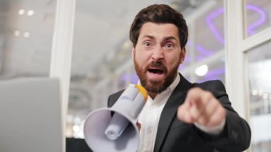 Kızgın işadamı megafona bağırır ve kamerayı işaret eder. Adam ofis ortamında masa başında oturuyor. Resmi giysiler içinde sinirli bir erkek megafonla konuşuyor ve el hareketi yapıyor.. .