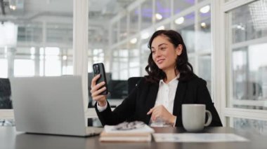 Takım elbiseli çekici bir iş kadınının ofiste bir video görüşmesi var. Kadın akıllı telefonuna bakar ve iş arkadaşları ya da müşterileriyle konuşur. Arka planda modern ofis alanı.