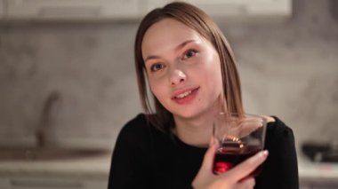 Güzel bir kadın sıcak bir mutfakta şaraptan hoşlanır. Zarif siyah elbiseli güzel bir kadın kutlama yapıyor, şarap içiyor ve gülümsüyor..