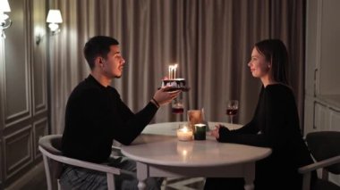 Romantik doğum günü kutlaması. Siyah elbiseli çekici bir adam ve kadın mutfakta bir masada oturuyorlar. Yanan mumlar ve lezzetli bir doğum günü pastası rahat bir ortam yaratır..