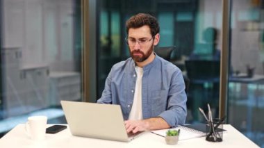 Günlük giysiler ve gözlüklerle odaklanmış iş adamı modern ofiste dizüstü bilgisayarla çalışıyor. İşyerindeki klavyeye bulanık arka planda konsantre olmuş bir adam. Not defteri ve akıllı telefon beyaz masada..