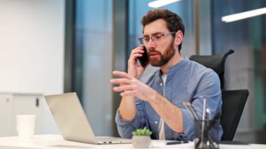 Gözlüklü ve rahat gömlekli odaklanmış işadamı cep telefonuyla modern ofiste çalışıyor. Adam ciddi ve meşgul hissediyor. Nişanlı erkek odaklanmış ve profesyonel görünüyor, iş kararları.