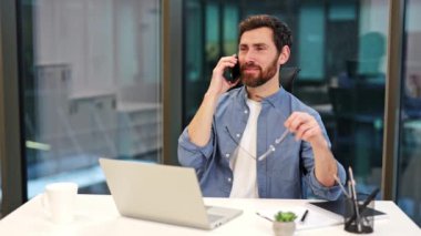 Başarılı iş adamı telefonda konuşuyor, gülümsüyor. Modern ofiste çalışan profesyonel bir erkek dizüstü bilgisayar kullanıyor, iş işlerini odaklı yönetiyor ve olumlu, mutlu bir tutum sergiliyor..