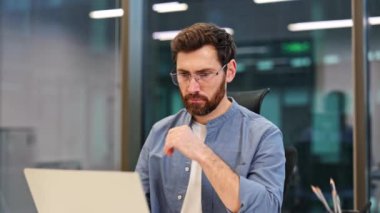 Gözlüklü nişanlı adam modern ofiste dizüstü bilgisayarda çalışıyor. İş yerindeki bilgisayarda günlük kıyafet giyen profesyonel işçiler. Düşünceli işadamı bütçe planlaması ve harcama optimizasyonunu analiz ediyor