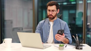 Modern ofiste dizüstü bilgisayar ve akıllı telefonla çalışan mavi gömlekli profesyonel bir erkek bilişim uzmanı. Hırslı iş adamı çoklu görev, görevlere odaklanmış ve önemli kararlar almış. İş, teknoloji