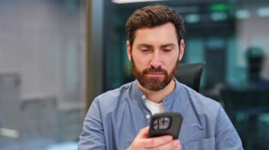 Gündelik gömlekli mutlu iş adamı modern bir ofiste telefon kullanıyor. Kendini mutlu hissediyor, sosyal medyayı kontrol ediyor, ve mutlu bir anda kafasını kaldırırken geniş, sağlıklı bir gülümsemeyle gülümsüyor..