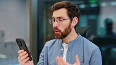 Ciddi bir iş adamı modern ofiste görüntülü görüşme yapıyor. Gözlüklü sakallı adam akıllı telefondan müşteriyle konuşuyor. İş, iletişim, teknoloji.