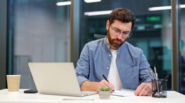 Gülümseyen iş adamı modern ofis masasında deftere yazıyor. Laptop, akıllı telefon, kahve, kalem ve kalem yakında. Meşgul profesyonel iş adamı şirket çalışanı, genç erkek işçi yazılım geliştiricisi