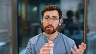 Ofisteki video görüşmesi sırasında kameraya konuşan, gözlüklü kendine güvenen iş adamı. Mavi gömlekli genç sakallı adam konuşuyor, anlatıyor, modern iş yerinde el kol hareketi yapıyor..