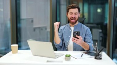 Modern bir ofiste telefonuna bakarken başarıyı kutlayan heyecanlı iş adamı. Heyecandan yumruğunu sallıyor, başarısını ve neşesini gösteriyor..