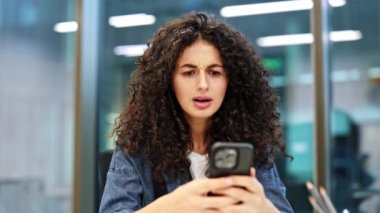 Sinirli kıvırcık saçlı iş kadını, modern ofisteki cep telefonundan gelen kötü haberlere tepki veriyor. Şok olmuş bir kadın, akıllı telefondan gelen olumsuz bilgilerle mesajı okur..