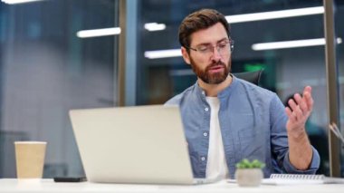 Odaklanmış işadamı, modern ofiste dizüstü bilgisayarla video görüşmesi yapıyor. Gözlük takan ve mavi gömlekli bir erkek çevrimiçi toplantıda notlar alıyor..