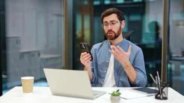 Odaklanmış iş adamı telefonda video görüşmesi yapıyor. Gözlüklü sakallı adam video bağlantısıyla konuşurken el kol hareketi yapıyor..