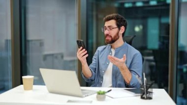 Gülümseyen iş adamı modern ofisteki akıllı telefondan video görüşmesi yapıyor. Genç erkek cep telefonuyla görüntülü konuşma yapıyor. İş, teknoloji, iletişim konsepti.