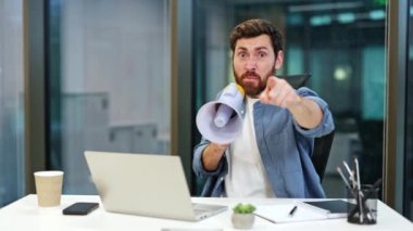 Kızgın işadamı megafona bağırıyor ve kamerayı işaret ediyor. Modern ofiste megafon kullanan öfkeli bir erkek. Sakallı adam ofiste bağırıyor..