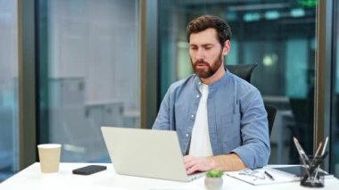 Sakallı yorgun işadamı masasında oturuyor, esniyor ve modern bir ofiste dizüstü bilgisayarıyla çalışıyor..