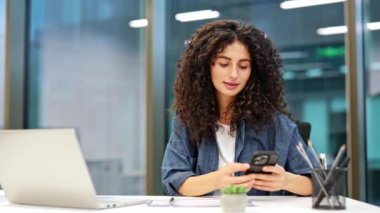 Modern ofiste dizüstü bilgisayar ve akıllı telefon kullanan kıvırcık saçlı genç bir iş kadını. İş yerinde çalışan meşgul kadın girişimci..
